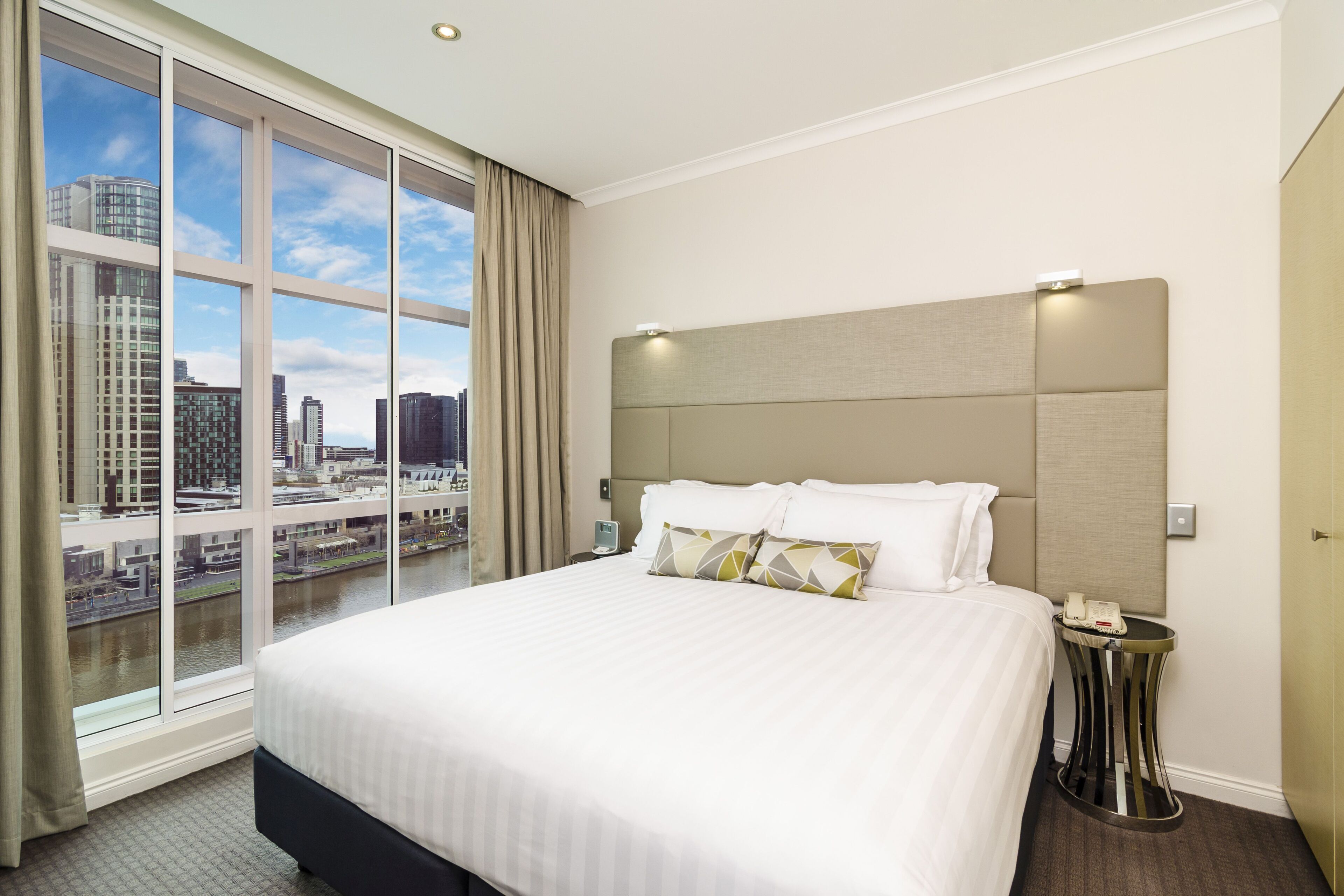 A hotel room with a large bed, a bedside table with a phone, and a view of a cityscape through floor-to-ceiling windows.