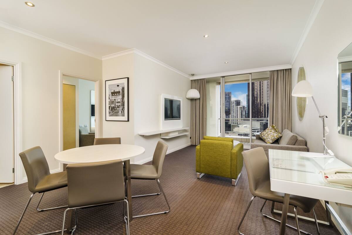 Une chambre d’hôtel avec une table ronde, des chaises, un canapé, une télévision, une lampe et une vue sur la ville.