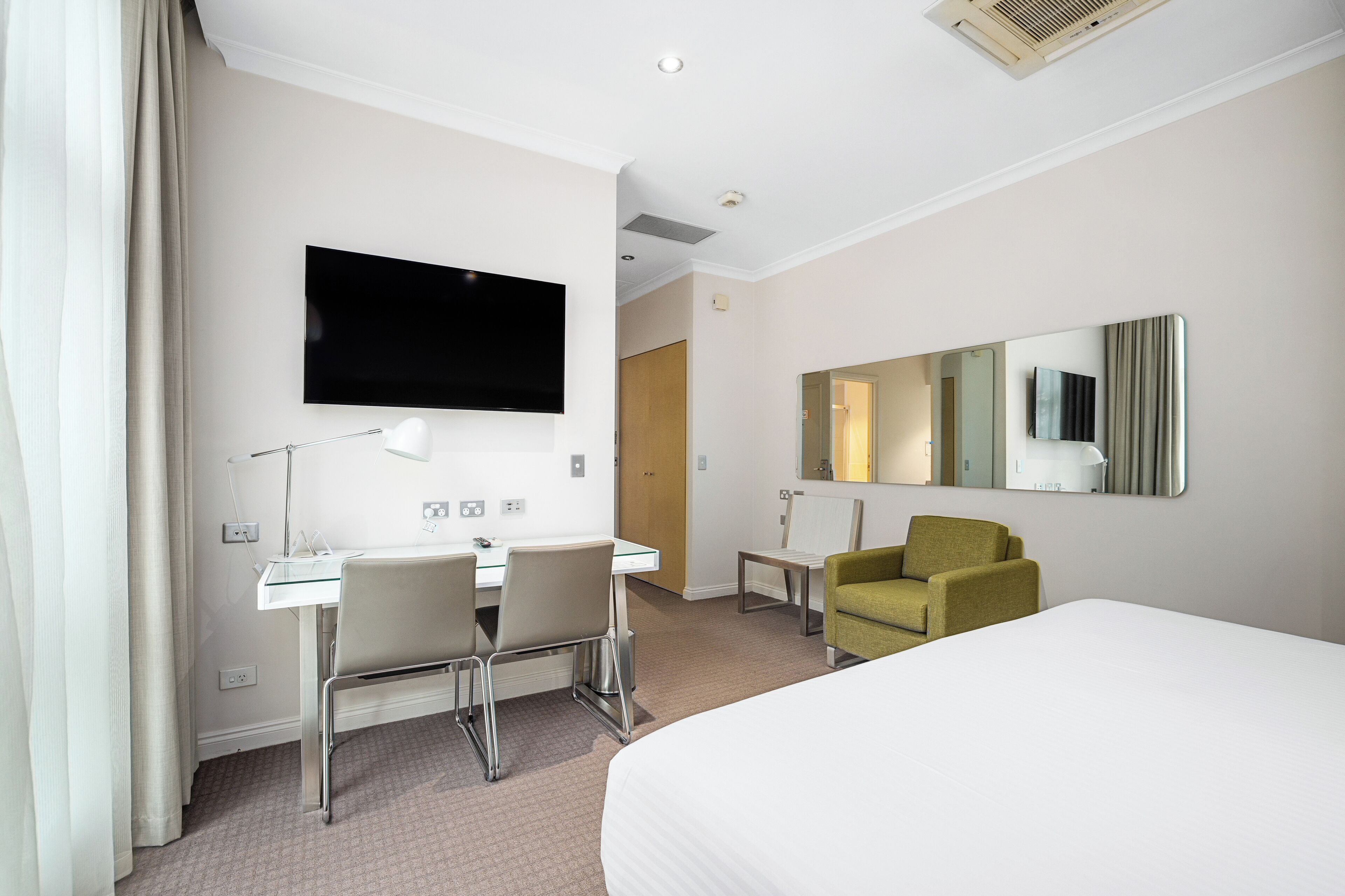 A hotel room with a bed, a desk with a chair, a television, a mirror, and a small table.