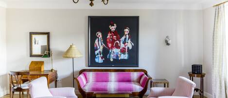 Un salon comprenant un canapé, deux fauteuils, une table basse en verre, un lustre, un tableau encadré, une lampe et une petite table.