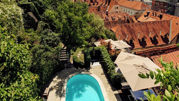 A swimming pool surrounded by lush greenery, with a view of historic buildings and a clear sky. - Schlossberghotel (Graz)