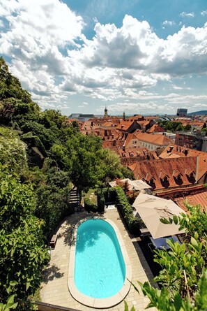 Une piscine entourée d’une végétation luxuriante, avec vue sur des bâtiments historiques et un ciel dégagé.