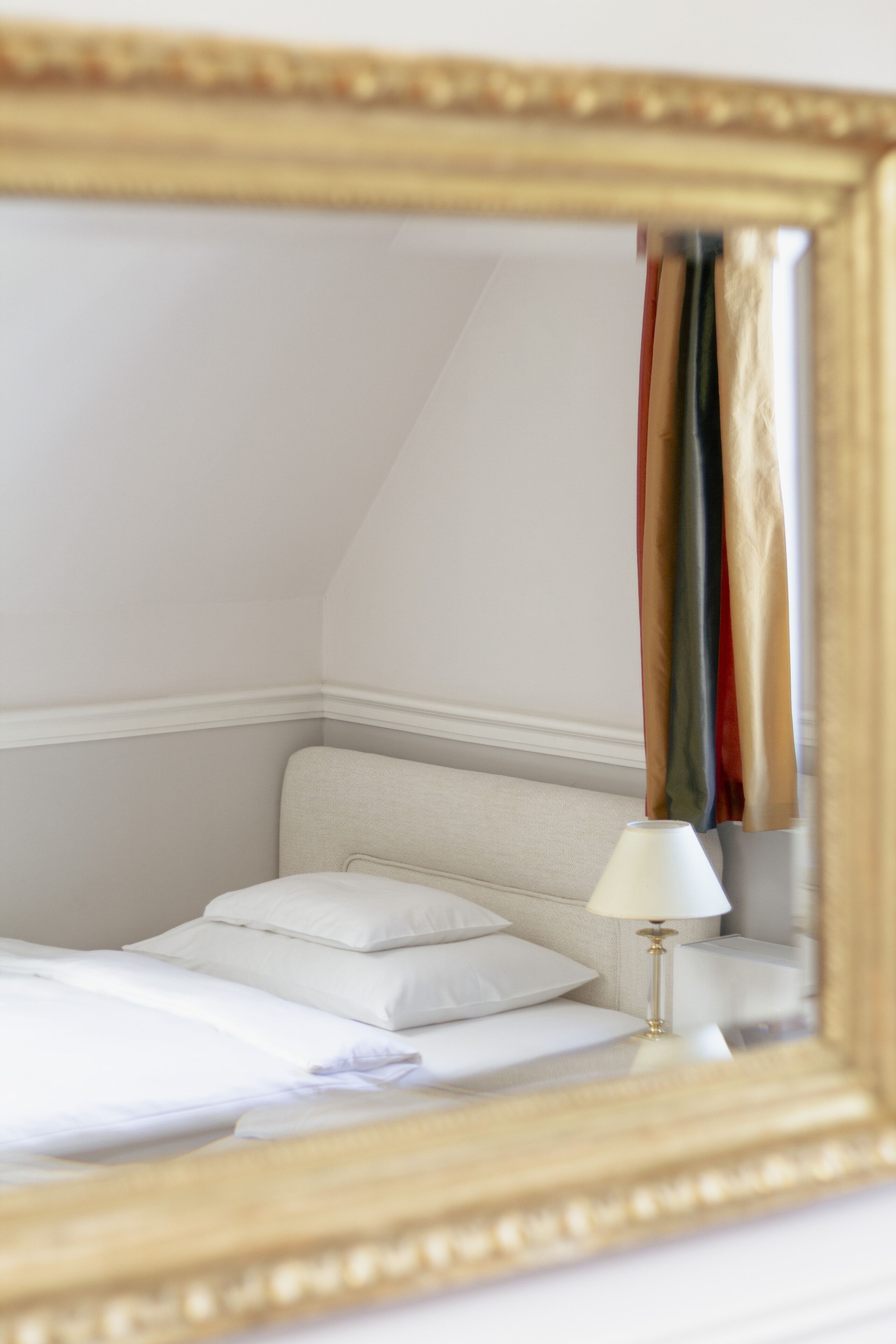A bedroom with a bed, white bedding, a bedside lamp, and a mirror reflecting a colorful curtain.