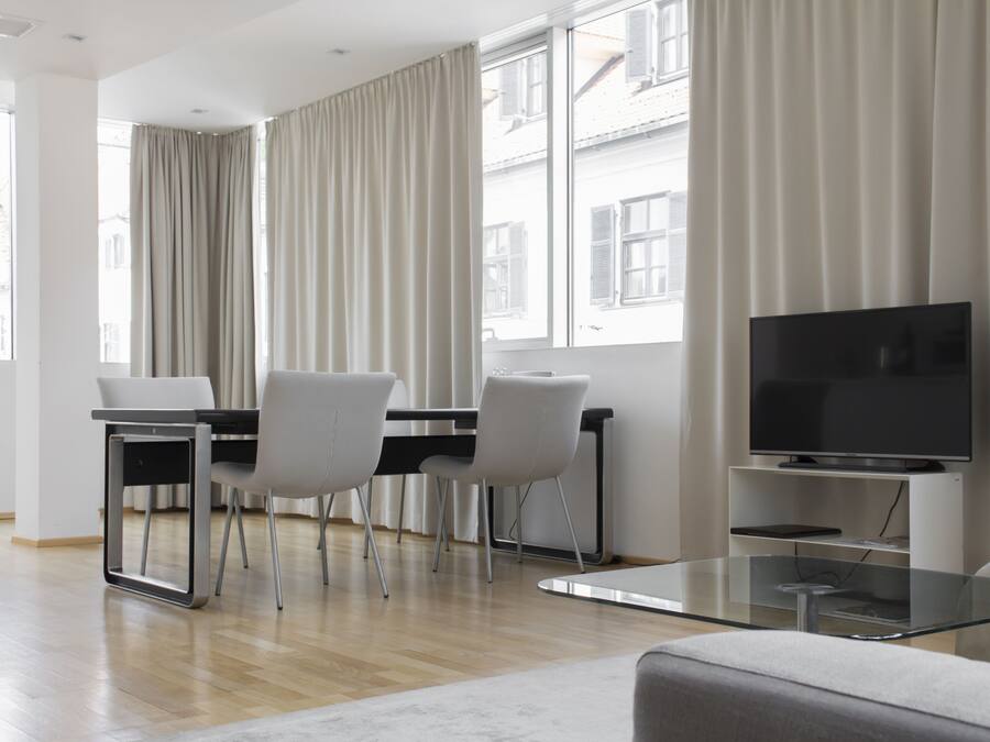 A modern living room with a glass coffee table, a flat-screen TV, a dining area with a table and chairs, and large windows with curtains.