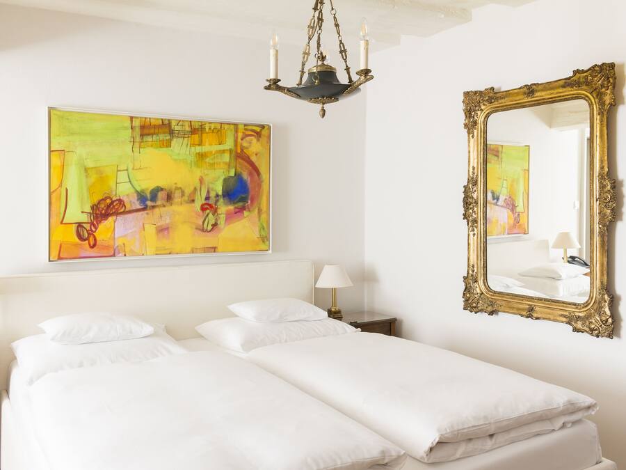 Habitación de hotel con una cama grande, una mesita de noche, una lámpara, un candelabro, un espejo y un cuadro abstracto de colores en la pared.