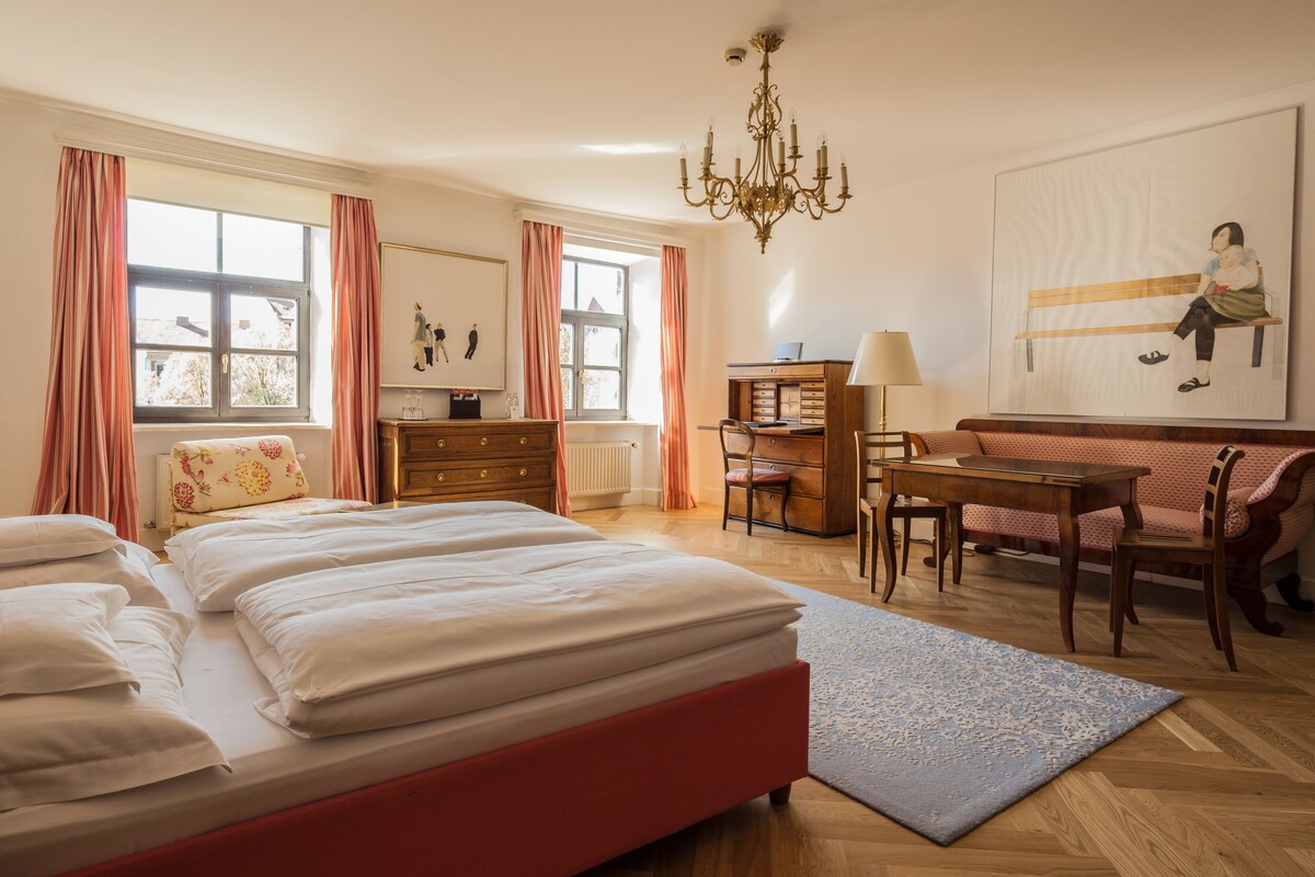 Ein Hotelzimmer mit Bett, Schreibtisch, Stuhl, Sofa, einer Kandelaber und Kunstwerken an der Wand.