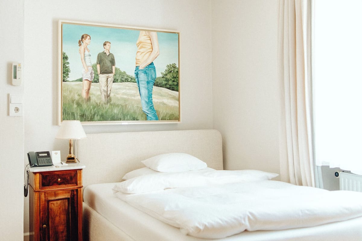Ein Schlafzimmer mit Bett, Nachttisch mit Lampe, einem Bild an der Wand und einem Fenster mit Vorhängen.