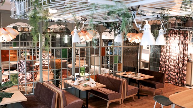 A restaurant with wooden tables and booths, hanging pendant lights, and a view through a glass partition. - Stadtoase Kolping Hotel (Linz)