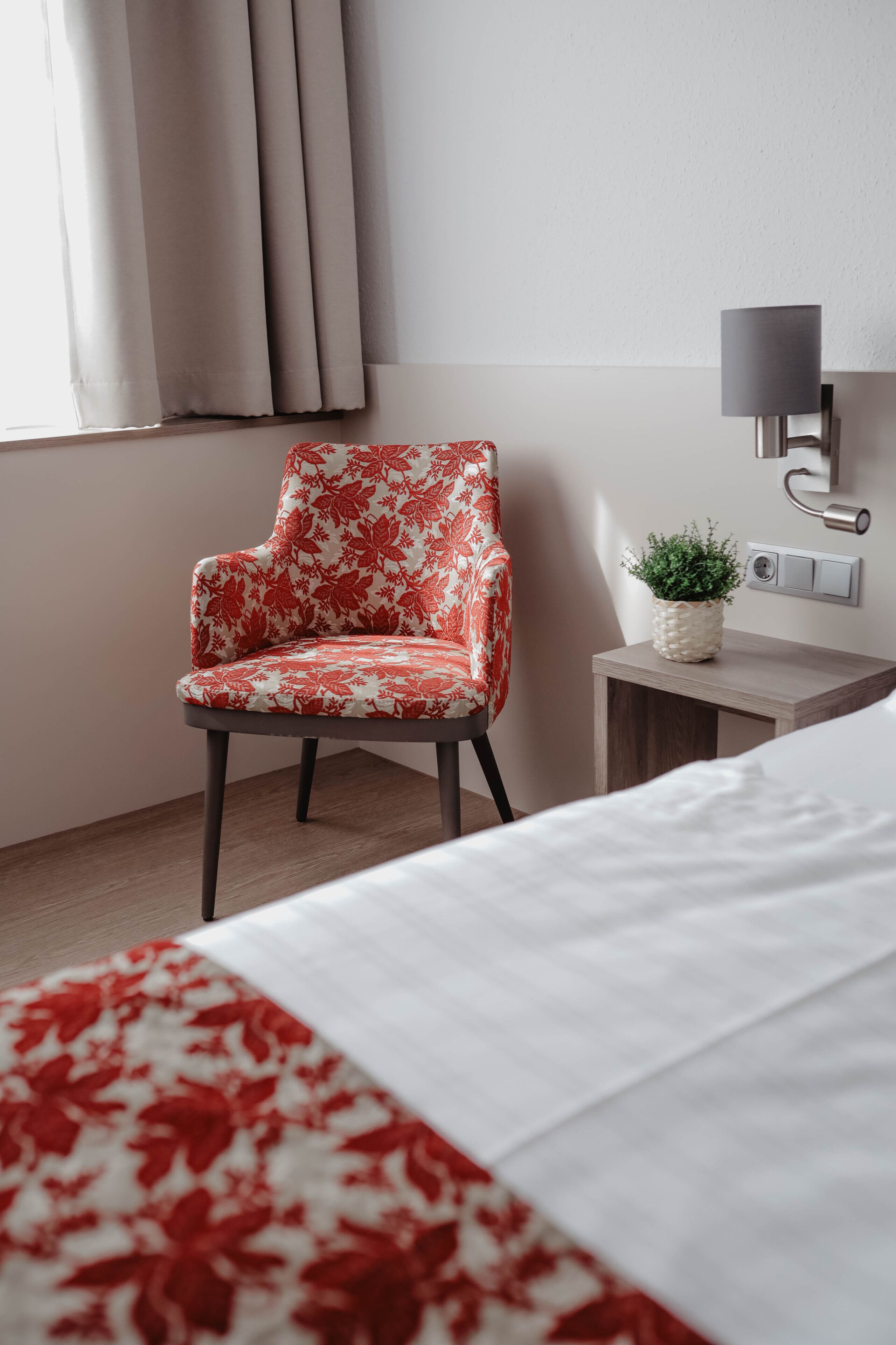 A hotel room with a floral-patterned chair, a bedside table with a plant, and a bed with a white and red blanket.