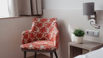A hotel room with a floral-patterned chair, a bedside table with a plant, and a bed with a white and red blanket.