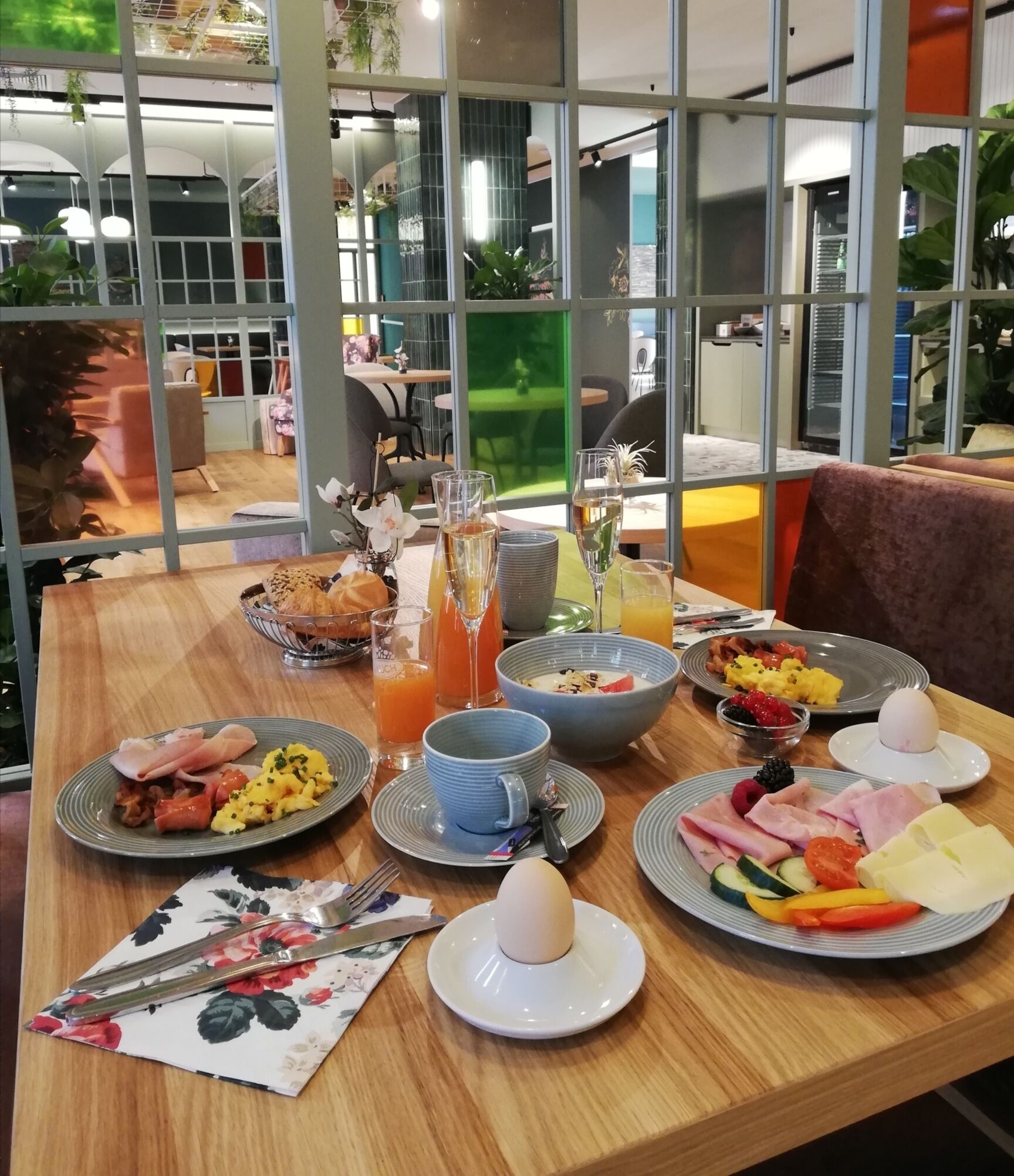 Una mesa de desayuno dispuesta con diferentes platos, entre ellos huevos, carnes y frutas, en un comedor moderno con ventanas amplias y plantas de interior.