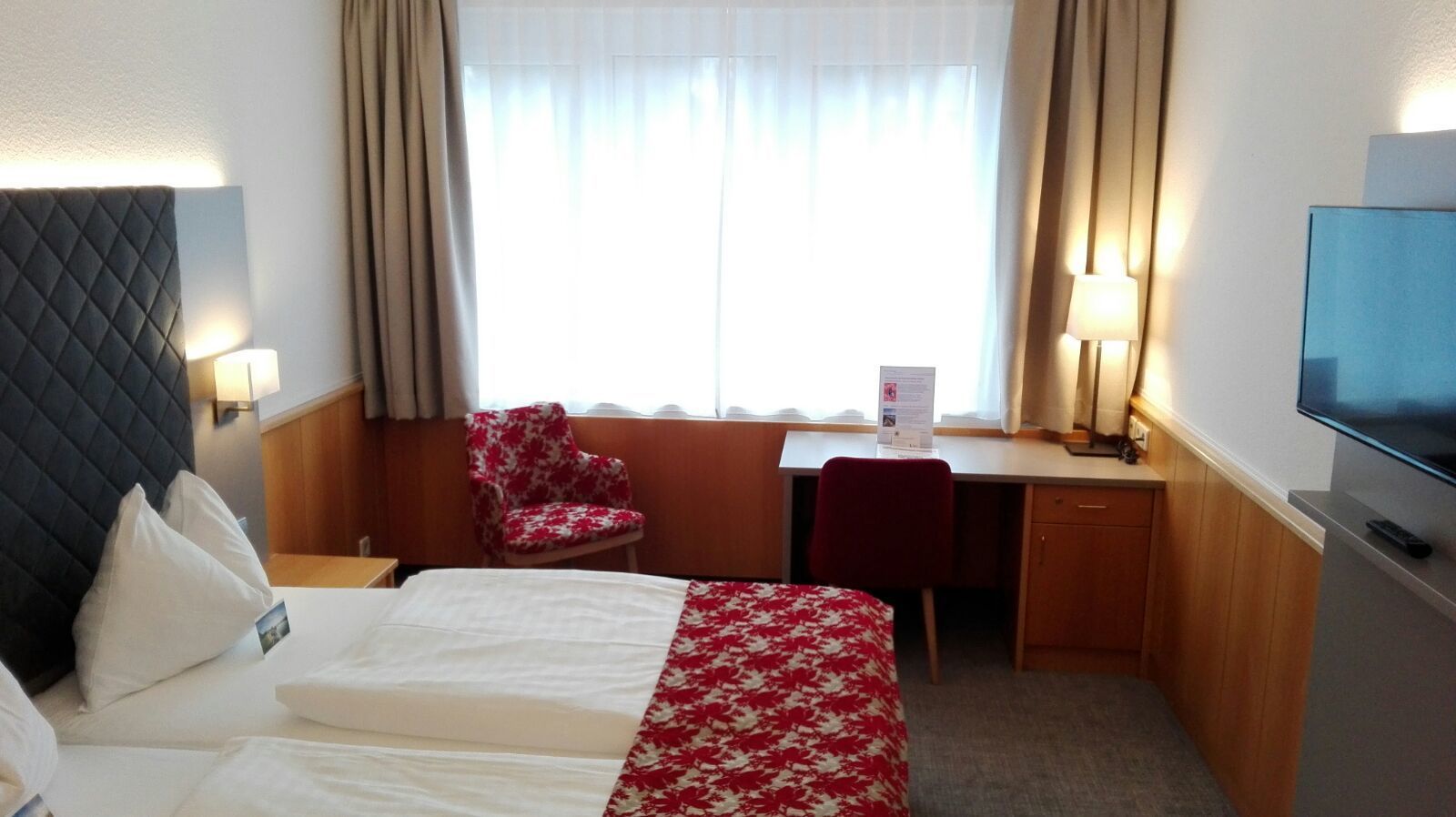 A hotel room with a bed, a desk, a chair, a TV, and a window with curtains.