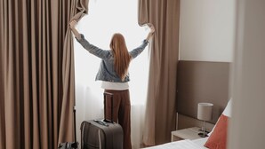 A hotel room with a bed, a suitcase, and curtains being adjusted by a person. - Stadtoase Kolping Hotel (Linz)