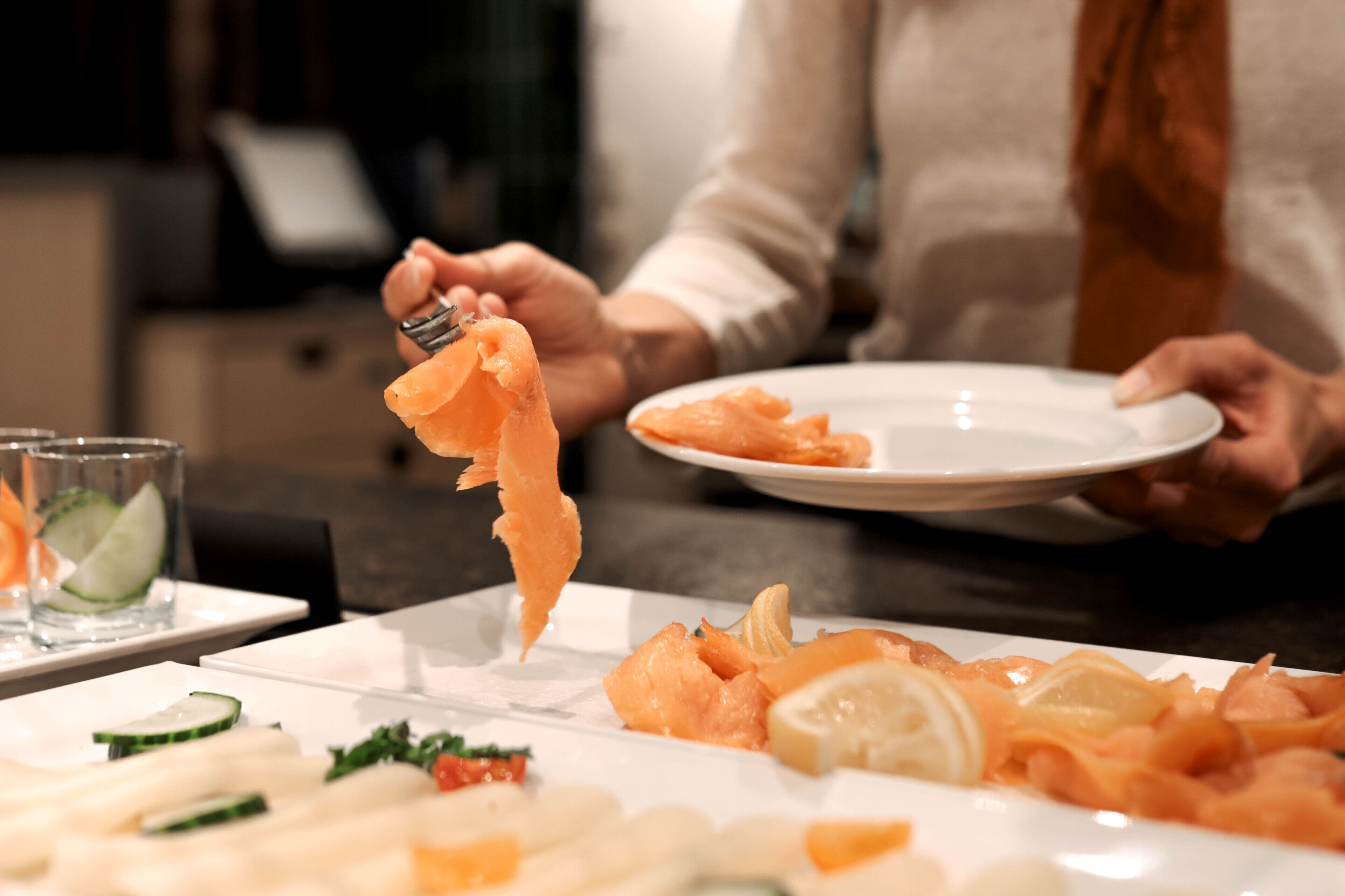 Eine Person hält eine Gabel mit einem Stück Lachs, ein Teller mit mehr Lachs und ein Glas mit einer Gurkenscheibe.