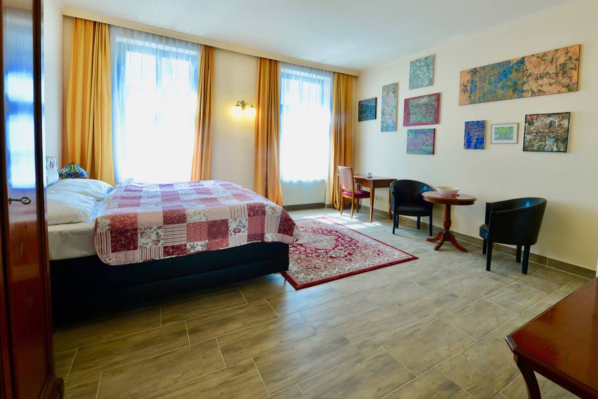 A hotel room with a bed, a desk, a chair, and a dining table. There are curtains on the windows and artwork on the walls.