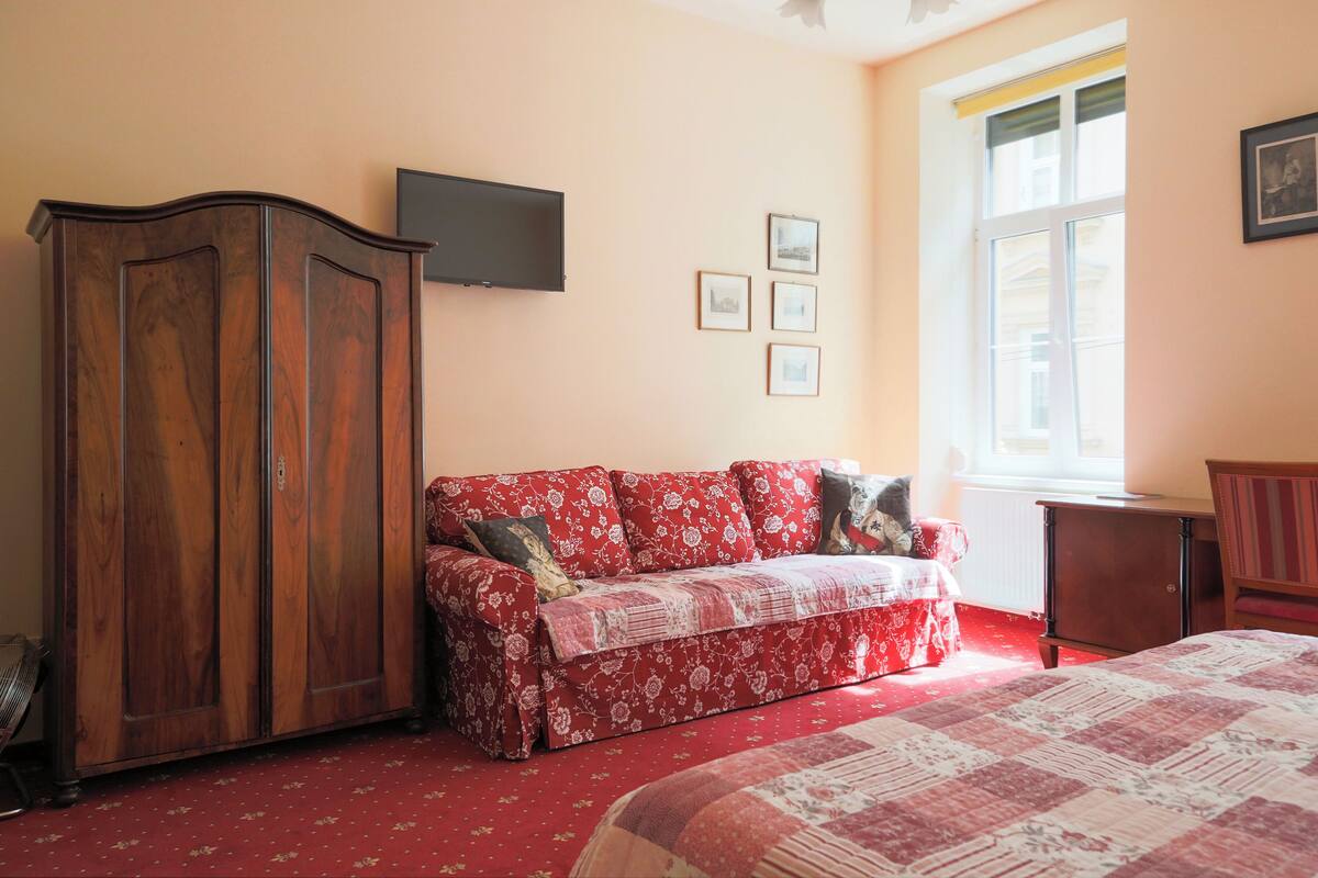 A bedroom with a wooden wardrobe, a red floral sofa, a bed with a quilt, a television on the wall, and a window with a view.