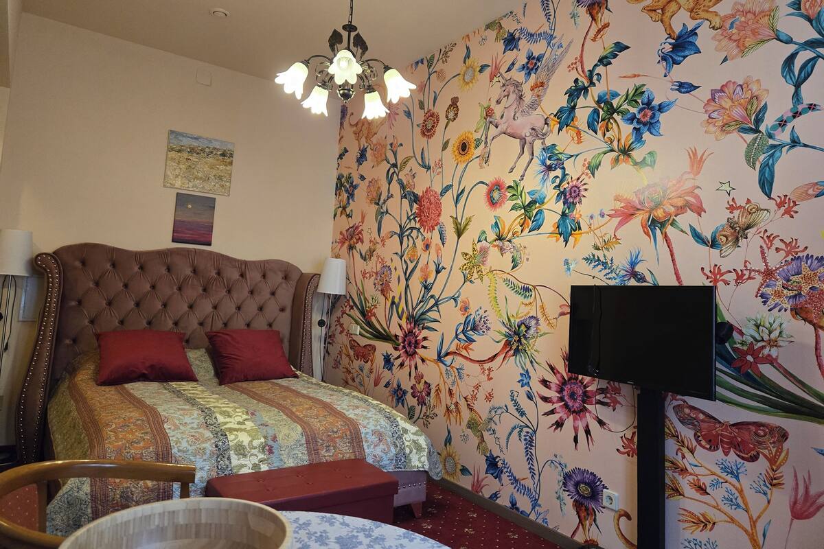 A bedroom with a tufted headboard, a large floral-patterned wall, a chandelier, and a television.