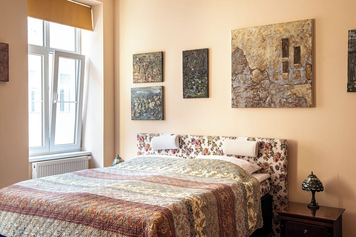 A bedroom with a floral bedspread, a large painting on the wall, and a bedside lamp.