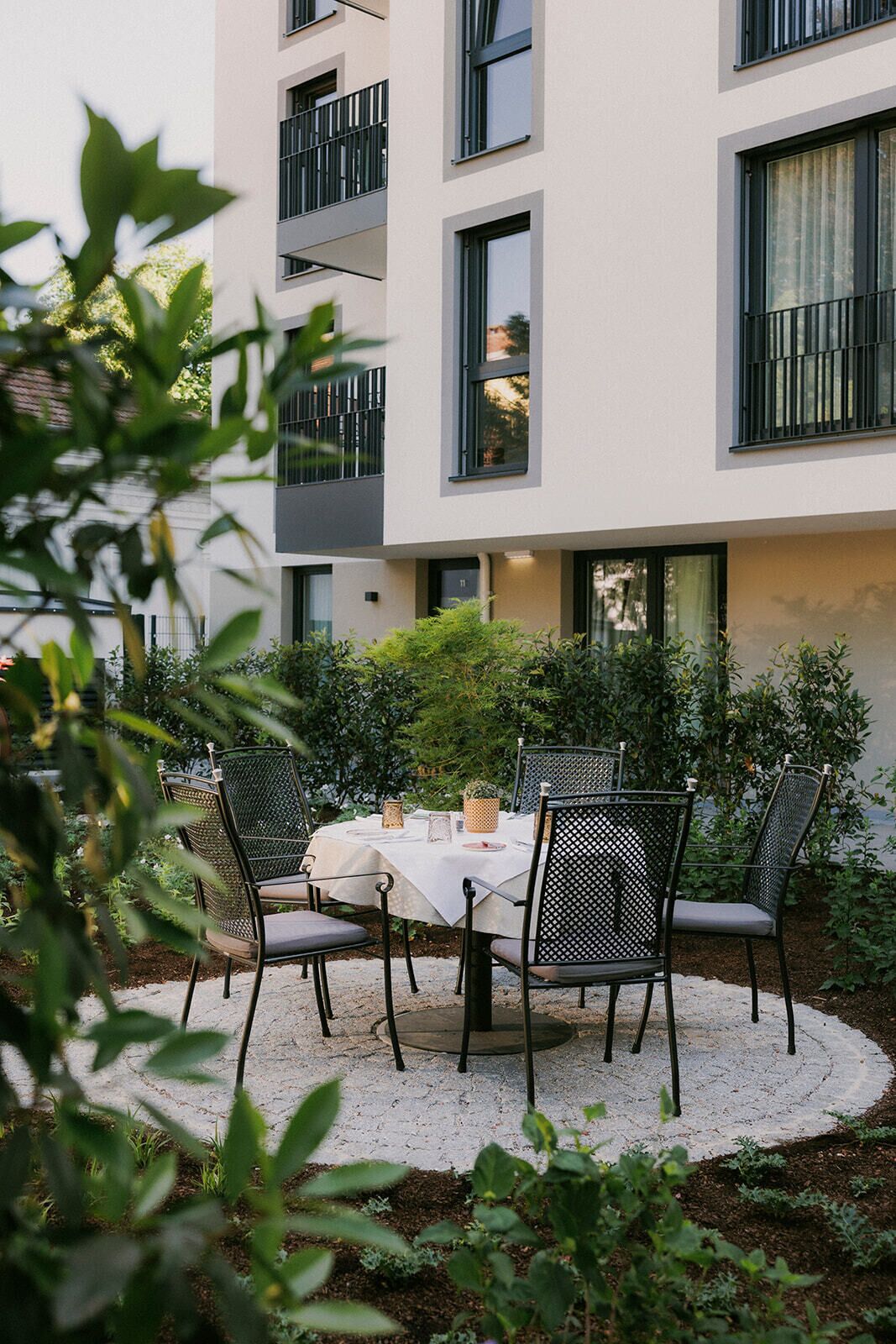 Un área de comedor al aire libre con una mesa redonda y cuatro sillas, rodeada de vegetación y un edificio con balcones.