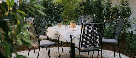 Un área de comedor al aire libre con una mesa redonda y cuatro sillas, rodeada de vegetación y un edificio con balcones.