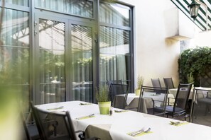 An outdoor dining area with tables and chairs, a potted plant, and a glass door leading to a garden.