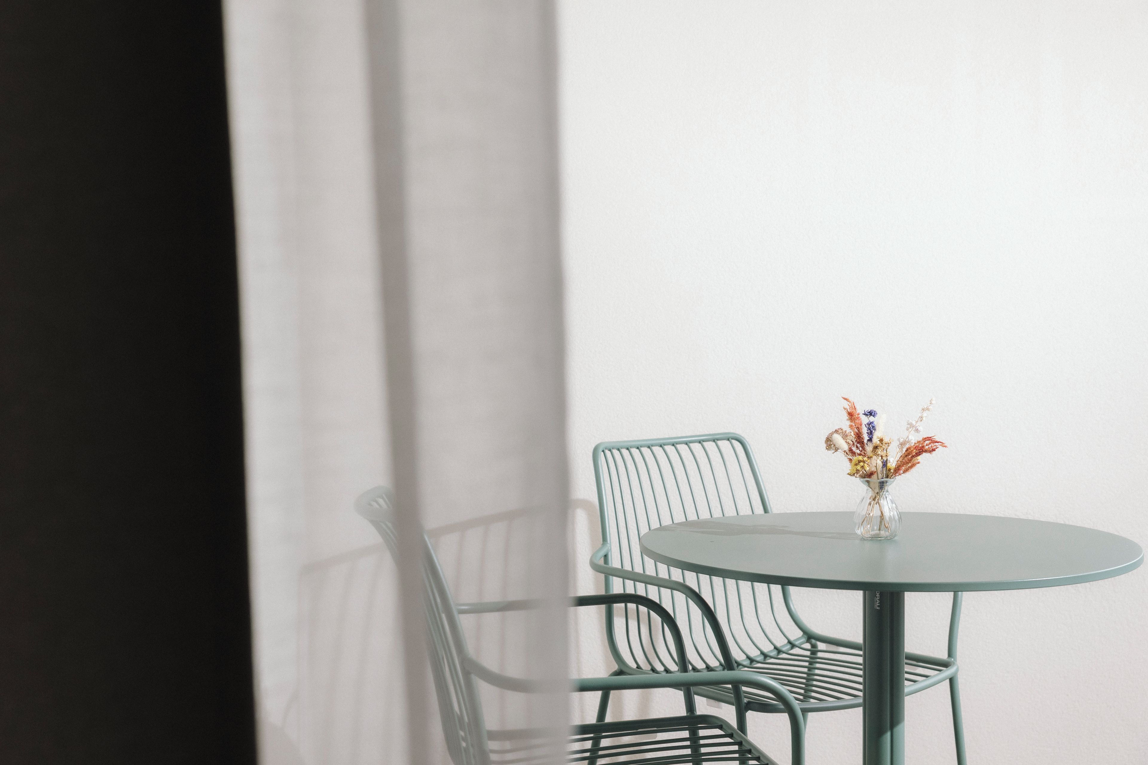 Una mesa de vidrio redonda con un florero, dos sillas de malla verde y una pared blanca.