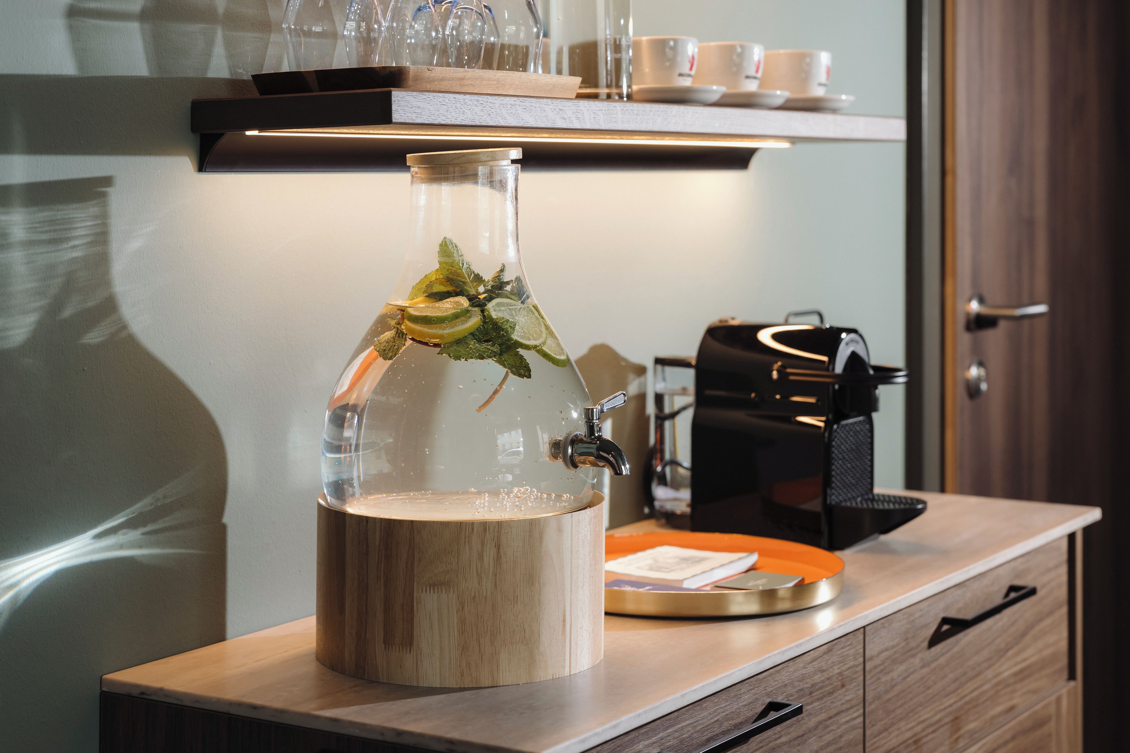 A glass decanter with a wooden base on a wooden countertop, a coffee machine, and a shelf with cups and glasses in the background.