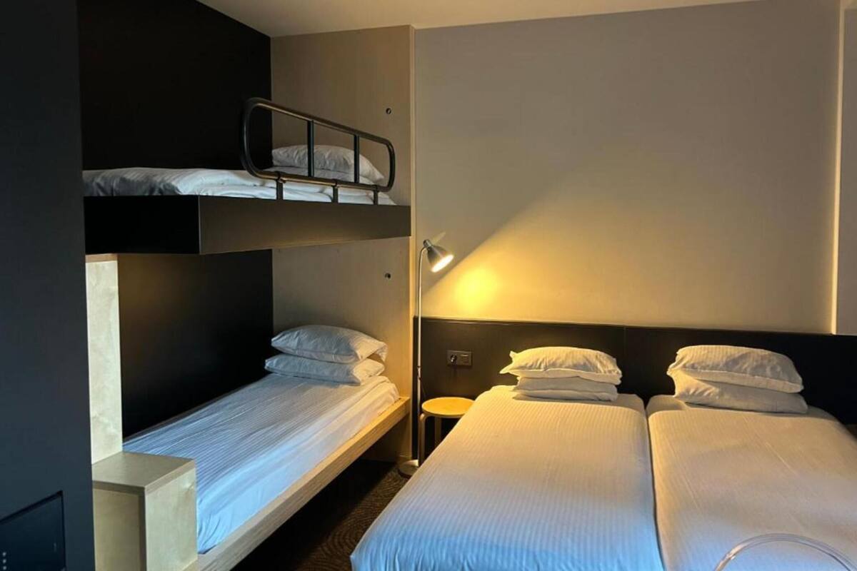 A hotel room with two single beds and a bunk bed, equipped with bedside tables and a lamp.