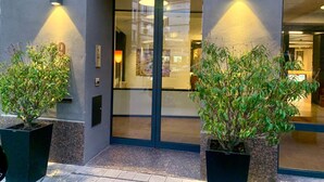 A modern building entrance with a glass door, two potted plants, and wall-mounted lights. - Hôtel Van Belle (Brussels)