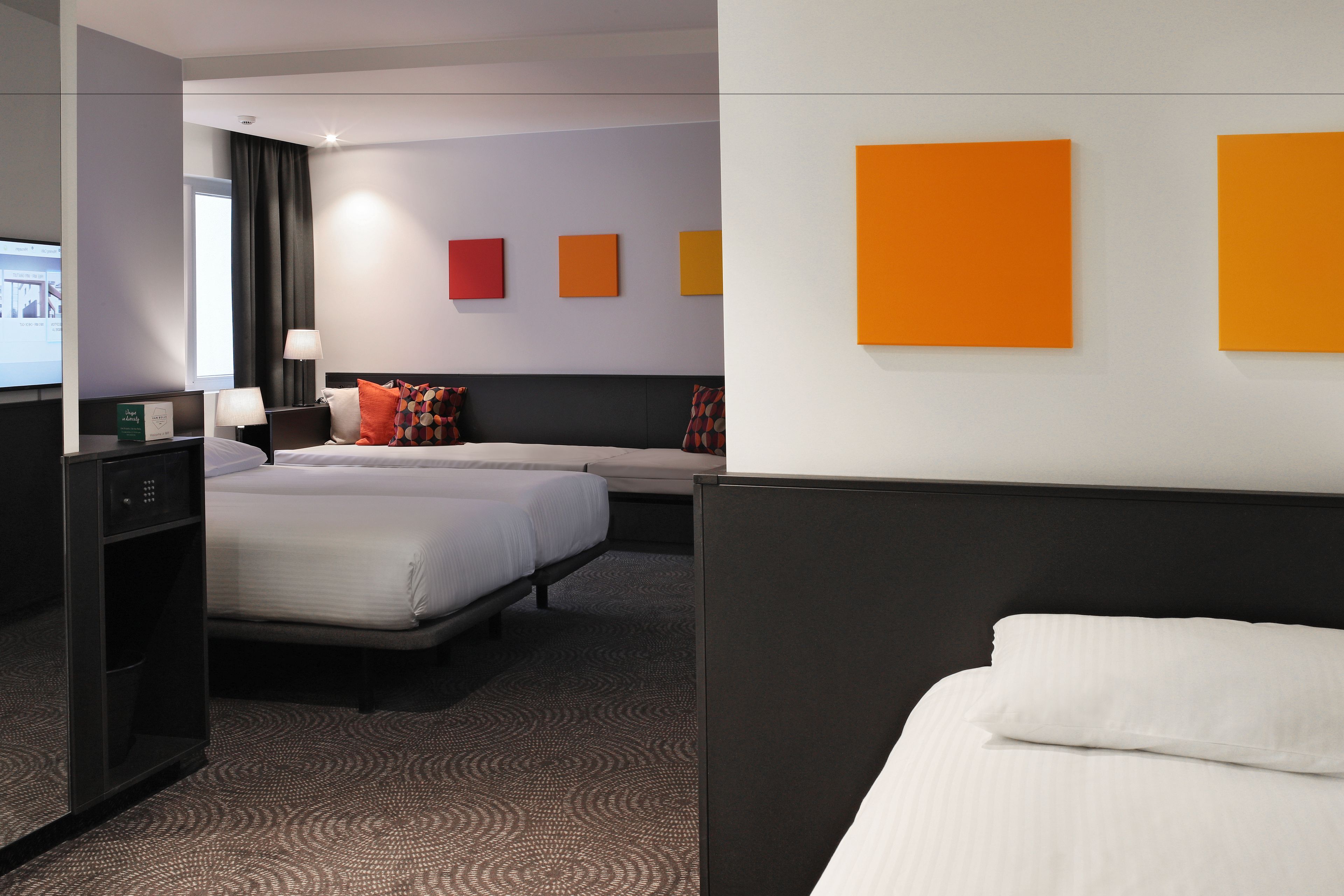 Hotelkamer met twee bedden, een bureau en een televisie. Op de muur hangen kleurrijke schilderijen en er ligt een kamerbreed tapijt met dessin.