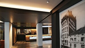 A hotel lobby with a reception desk, a bar area, and a large poster of a building with a sign reading "HOTEL RESTAURANT VAN BELLE". - Hôtel Van Belle (Brussels)