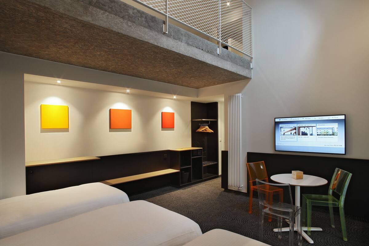 A hotel room with a bed, a bench, a small table with chairs, a TV screen, and a coat rack.