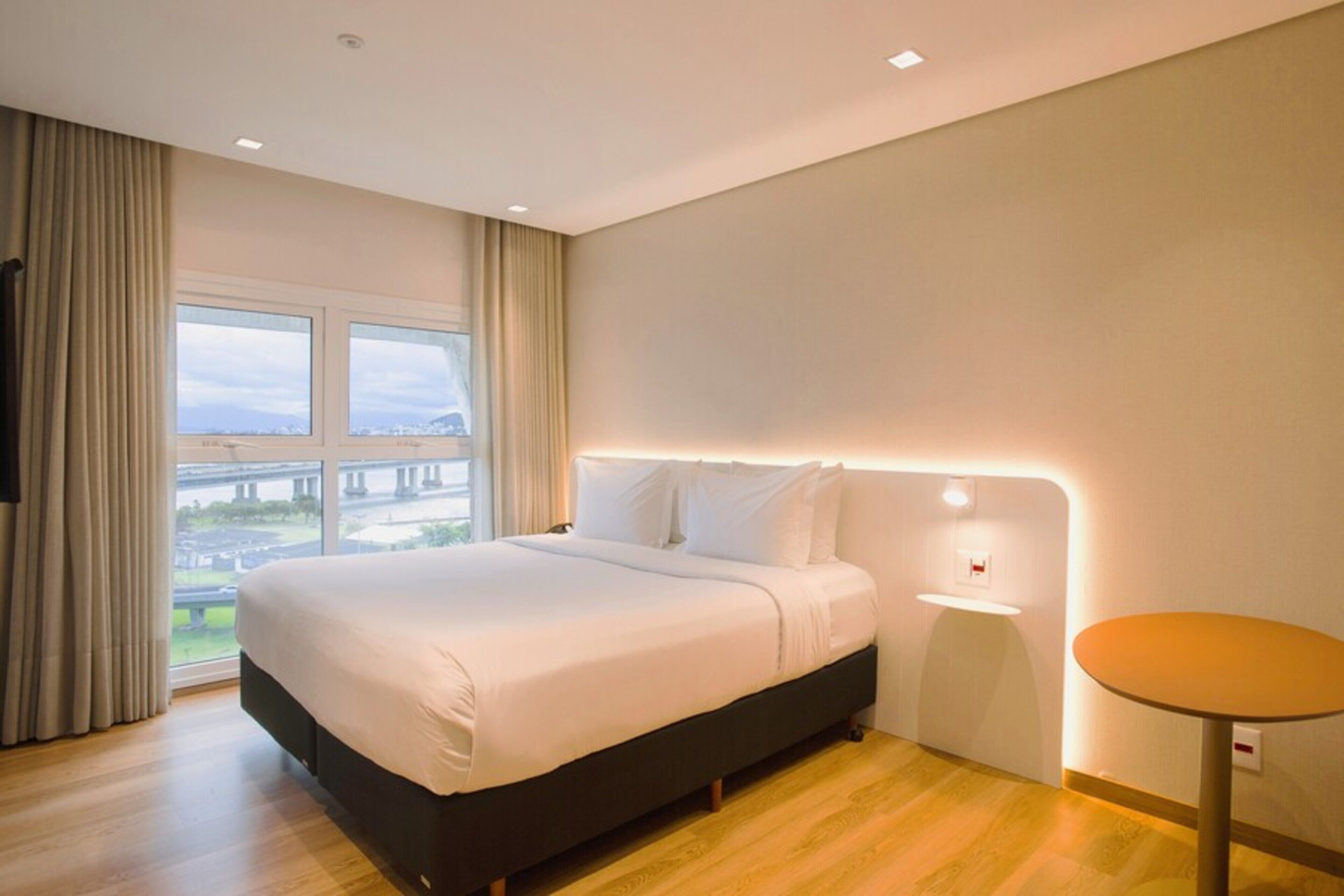 A modern hotel room with a large bed, a small bedside table, a round table, and a view of a bridge through the window.