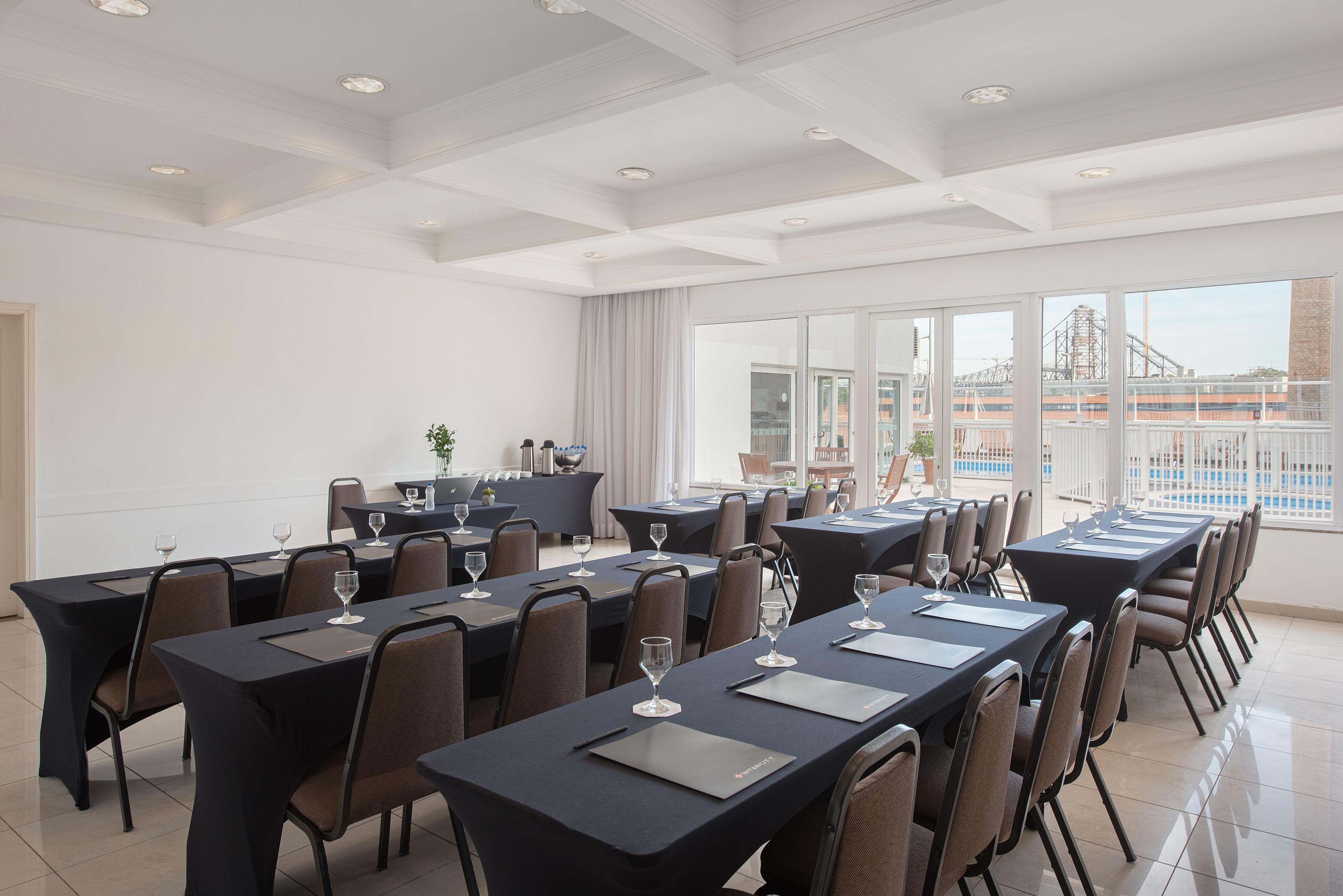 Une salle de conférence dotée de plusieurs tables, de chaises et de verres, avec de grandes fenêtres offrant une vue sur un pont et une piscine.