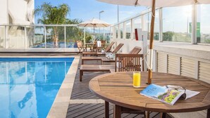 A poolside area with wooden lounge chairs, a round table with a magazine and a glass of orange juice, and large umbrellas providing shade. - Intercity Florianópolis (Florianópolis)