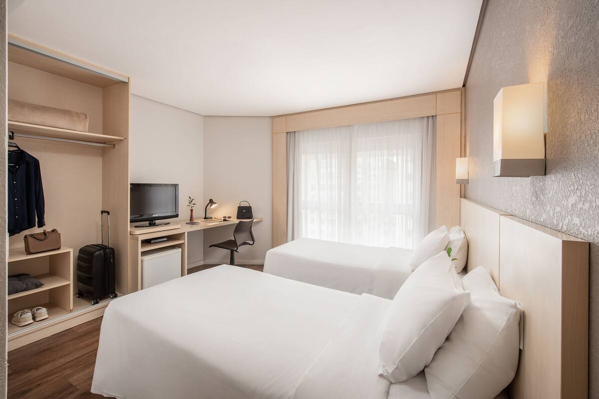 Habitación de hotel con dos camas, un escritorio con televisor, un armario y una lámpara de escritorio pequeña.
