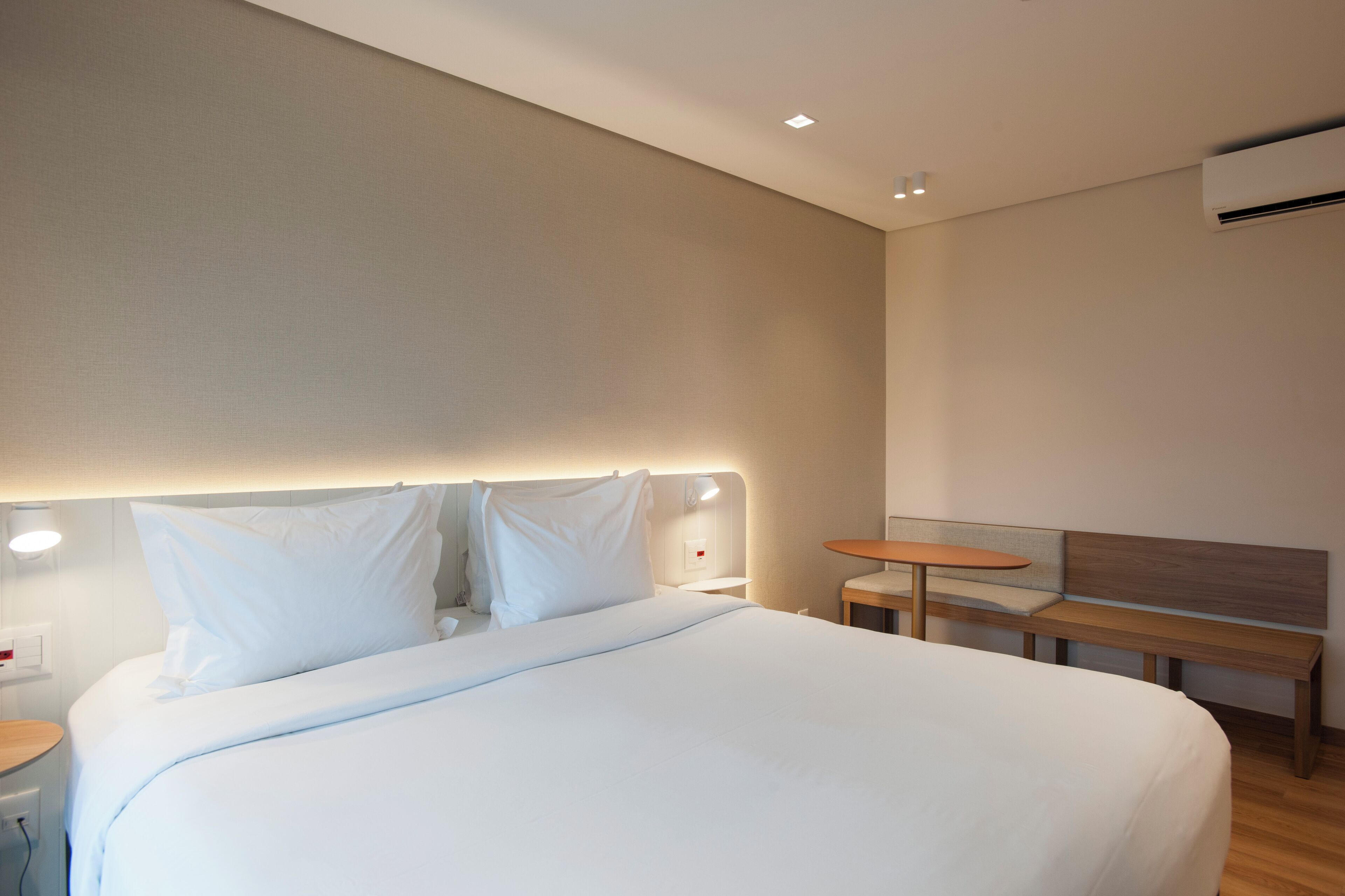 A modern hotel room with a large bed, a bench, a small table, and a wall-mounted air conditioner.
