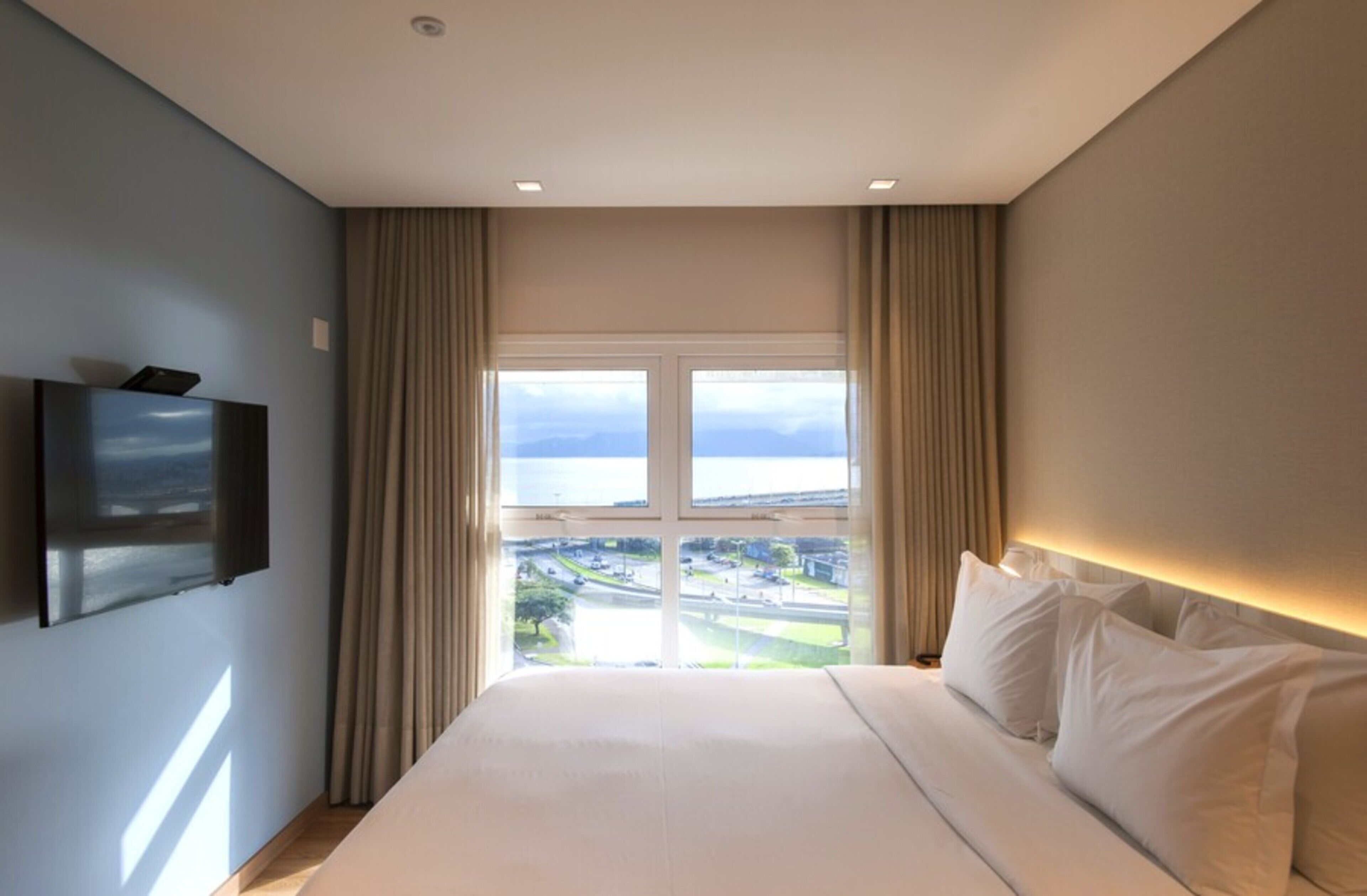 A hotel room with a large bed, a flat-screen TV mounted on the wall, and a window with a view of a cityscape.