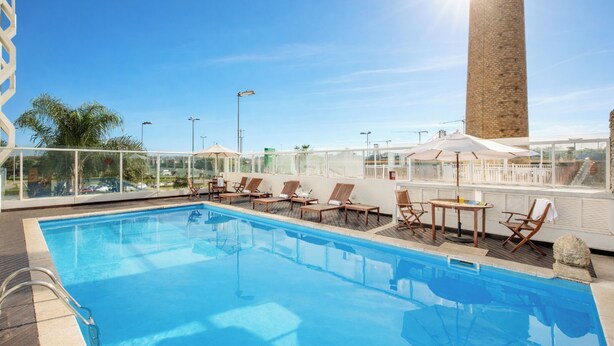 A rooftop pool with clear blue water, sun loungers, umbrellas, and a tall brick tower in the background.
