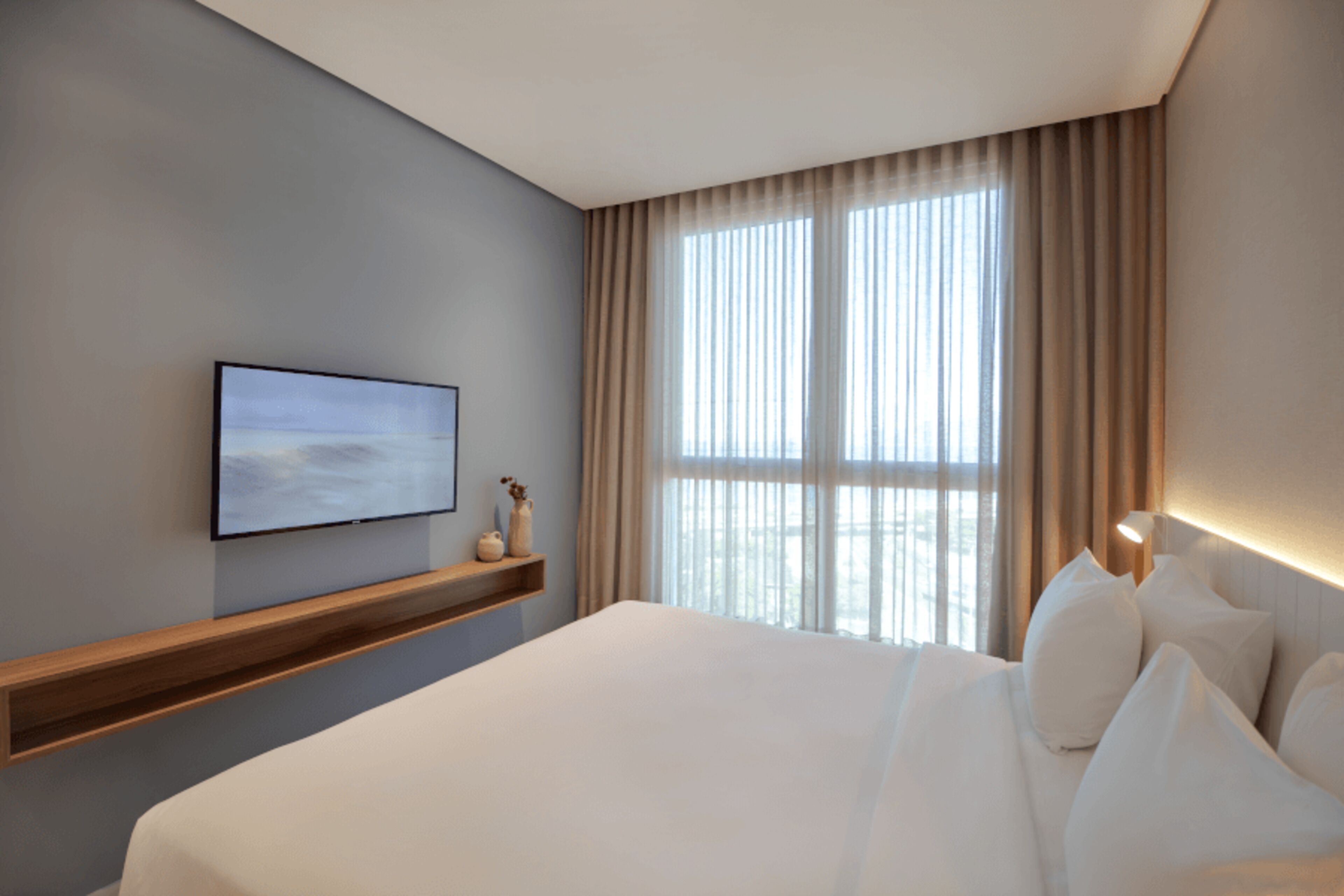A modern hotel room with a large bed, a mounted TV, a window with curtains, and a wooden shelf with decorative items.