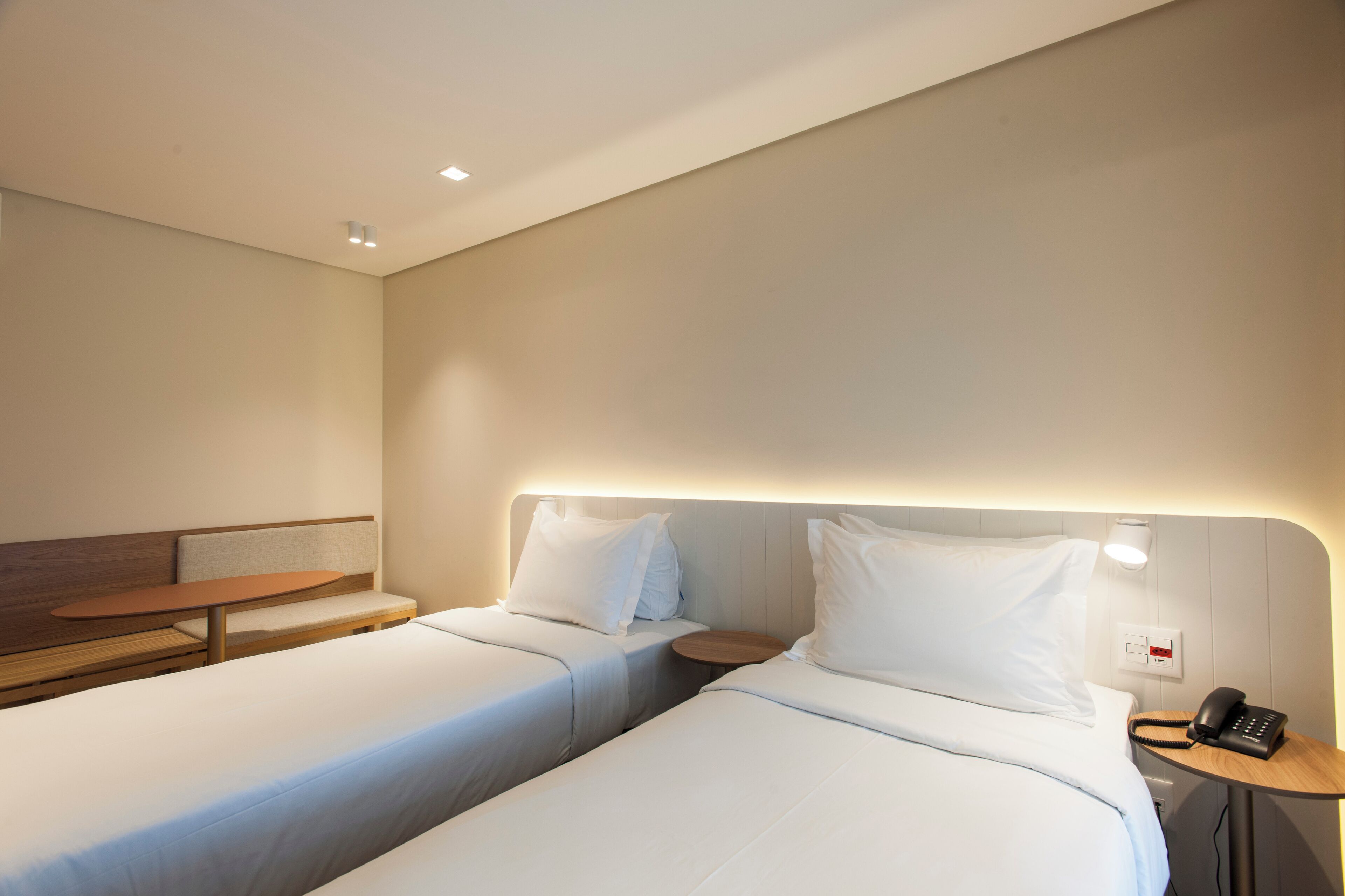 Una habitación de hotel con dos camas individuales, una mesita de madera, una mesita de noche con teléfono e iluminación en la pared.
