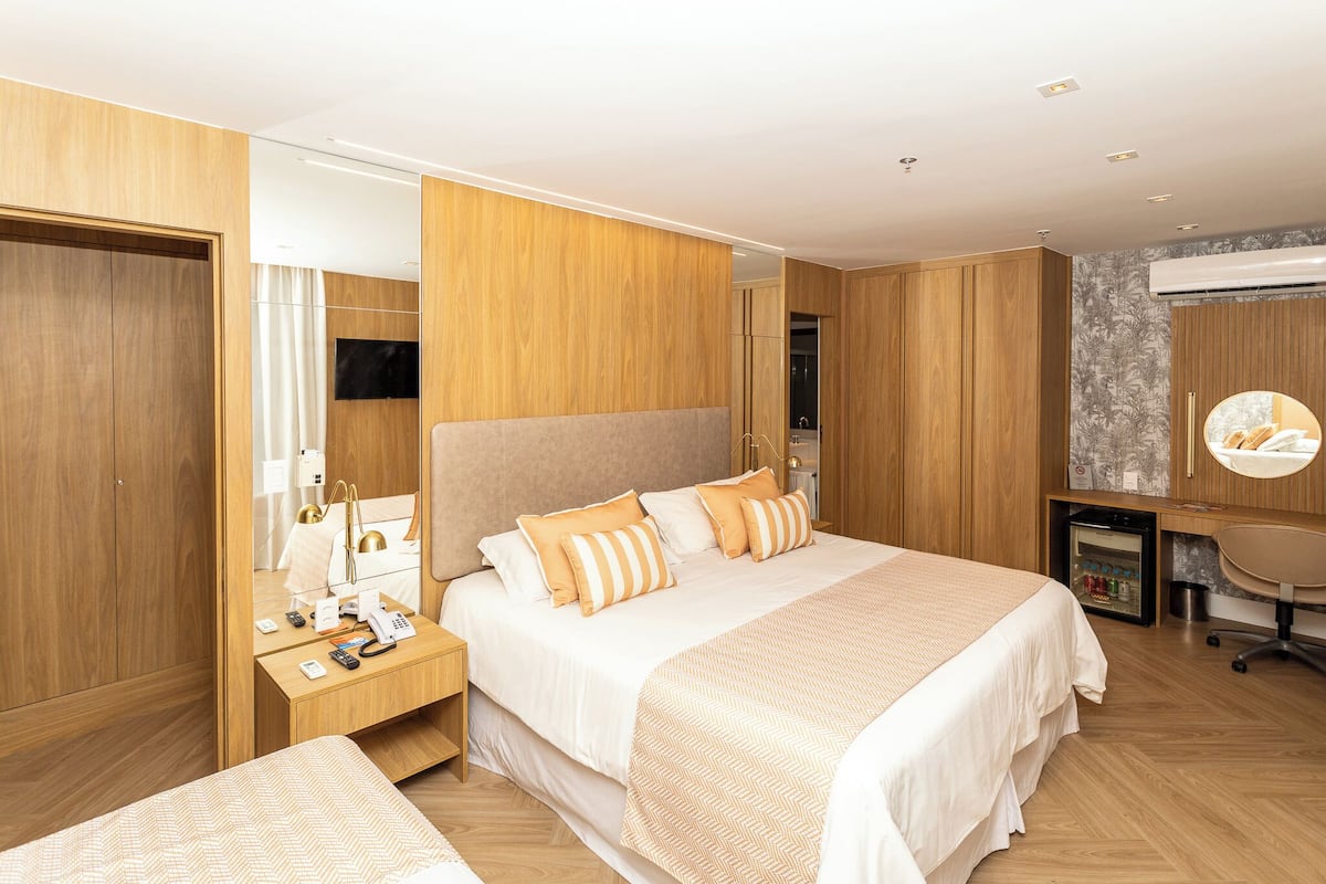 Habitación de hotel moderna con una cama grande, armario de madera, mesita de noche con lámpara, un pequeño escritorio con silla y aire acondicionado integrado.