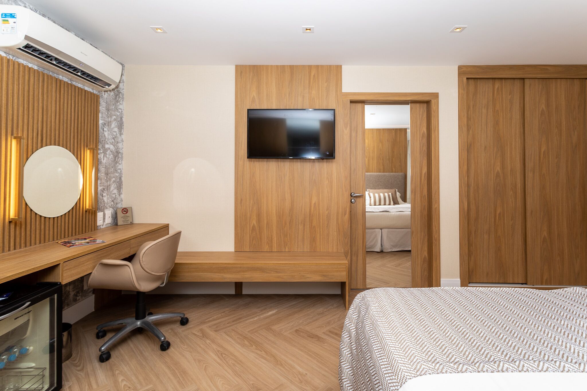 Habitación de hotel moderna con escritorio de madera, una silla cómoda, cama con ropa de cama estampada, televisor de pantalla plana montado en la pared y un armario.
