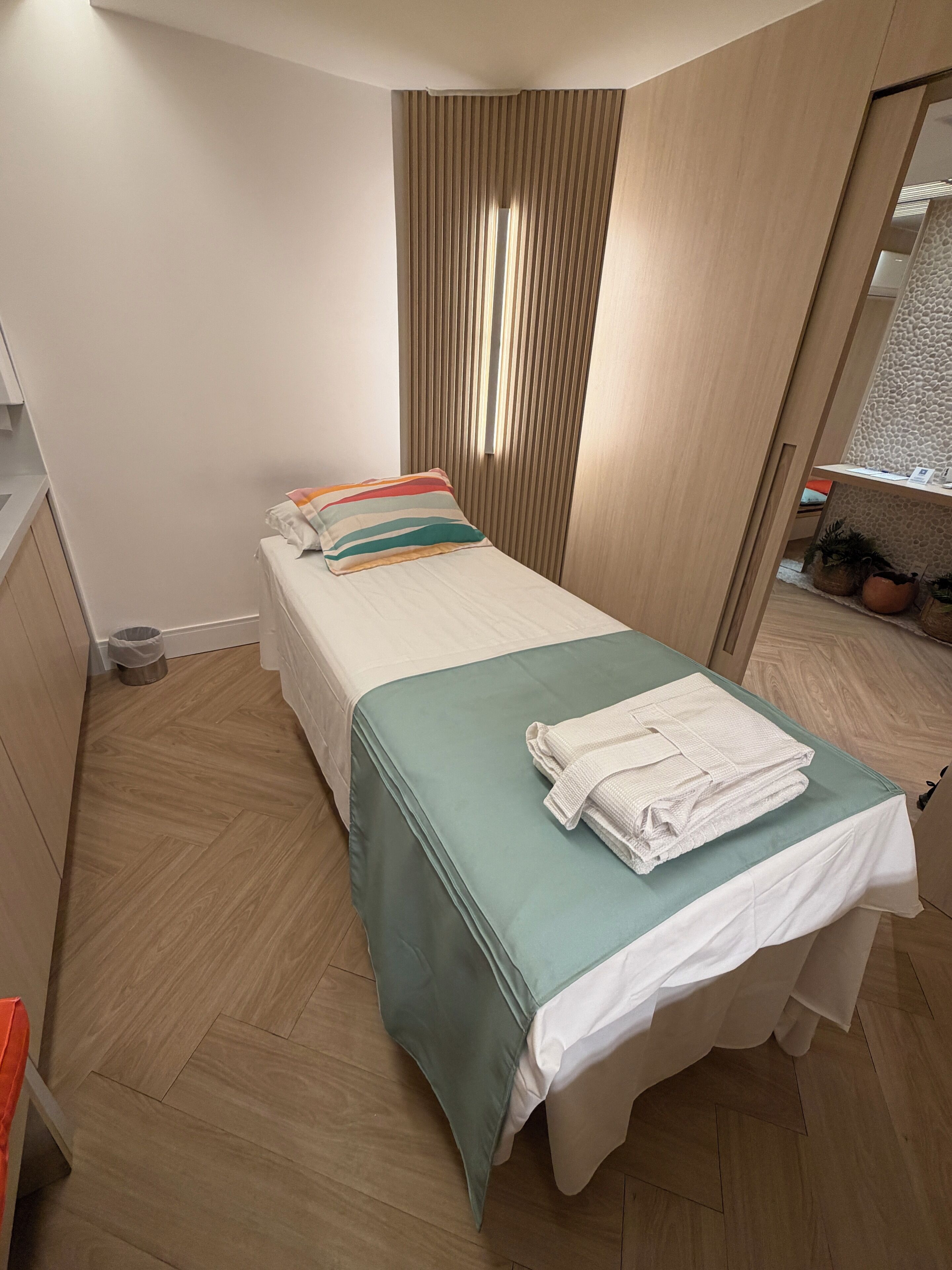 A small, neatly arranged bedroom with a single bed, a window with vertical blinds, a small table with a plant, and a trash bin.