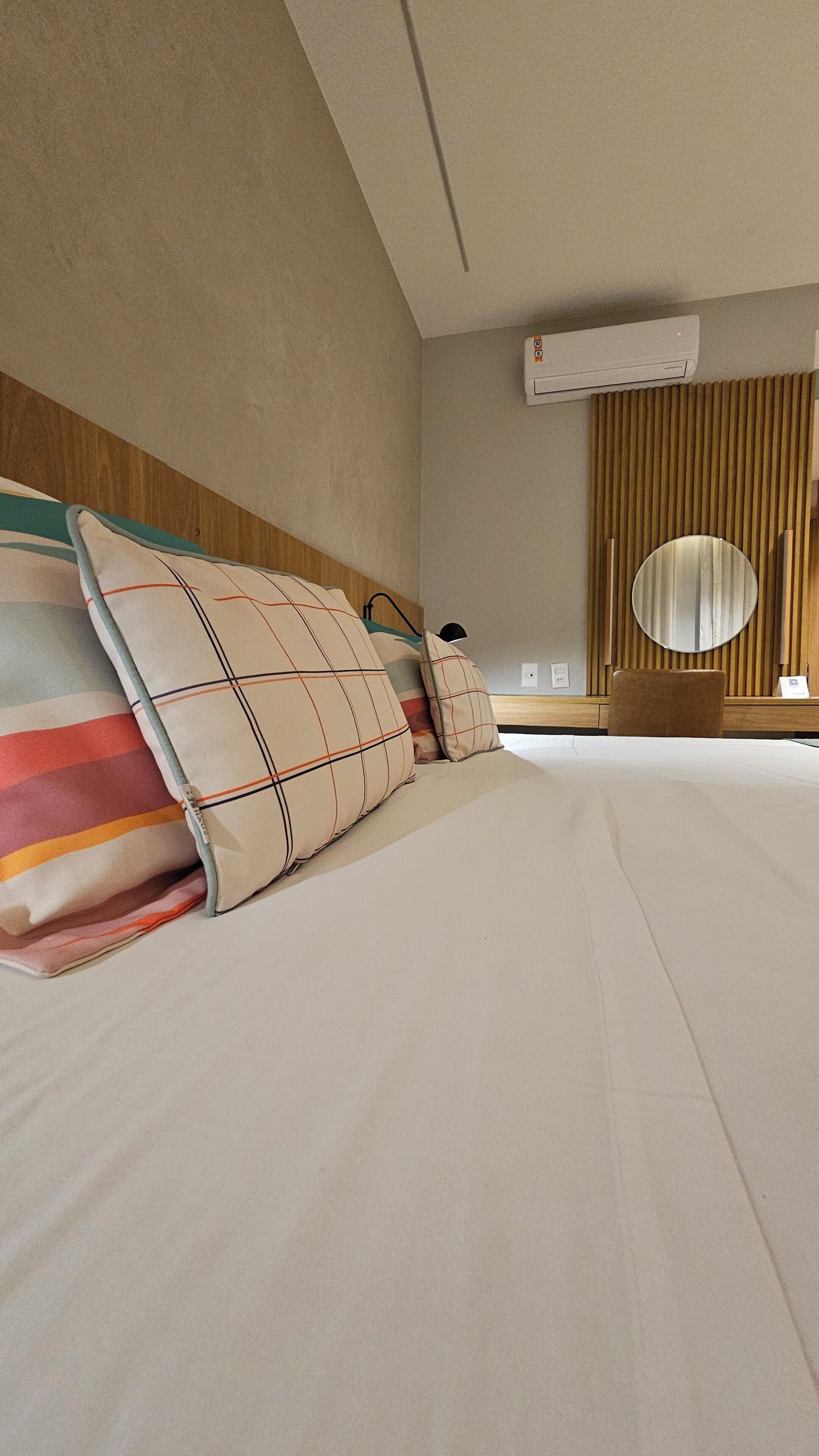 A hotel room with a bed, pillows, a round mirror, a wooden headboard, and an air conditioning unit.