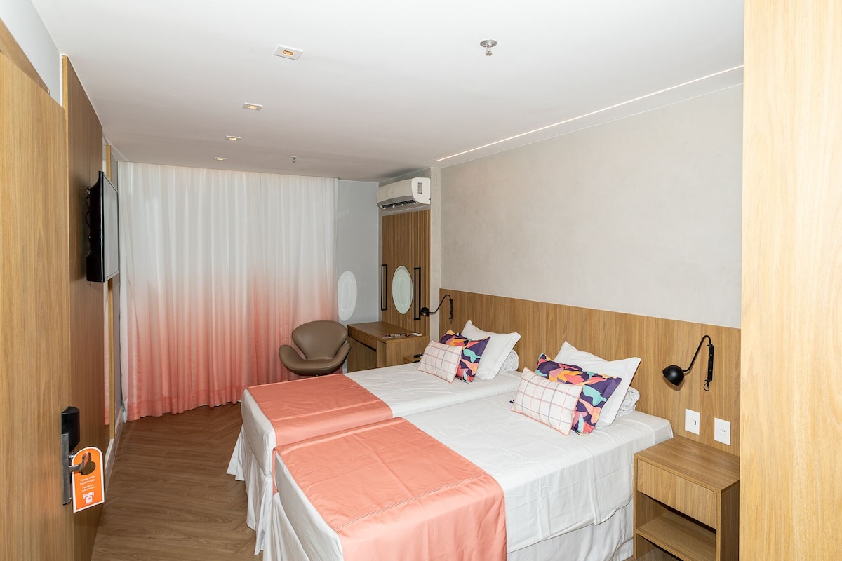 A hotel room with two beds, a desk, a chair, a TV, and an air conditioning unit.