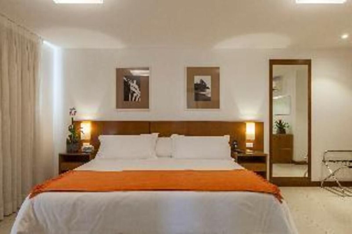 Una habitación de hotel con una cama, mesitas de noche, un espejo y obras de arte en la pared.