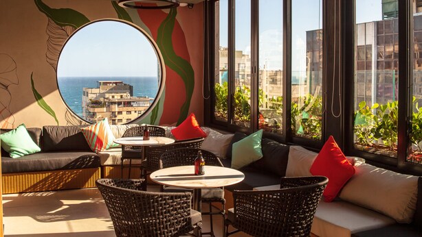 Un salón moderno con mobiliario de mimbre, una mesa redonda y un ventanal con vistas al mar y edificios.