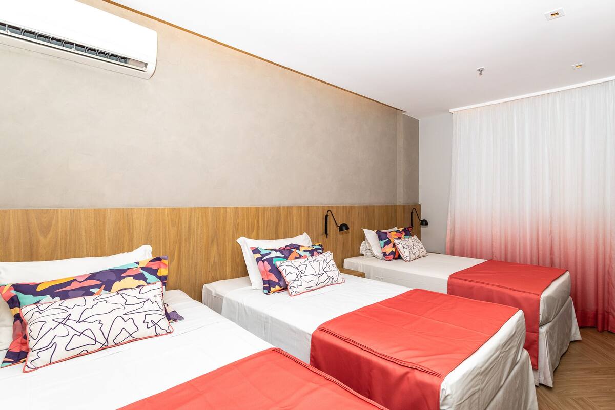 Habitación de hotel con tres camas, cada una con cubrecama rojo y almohadas estampadas, cabecera de madera, unidad de aire acondicionado y ventana con cortinas translúcidas.