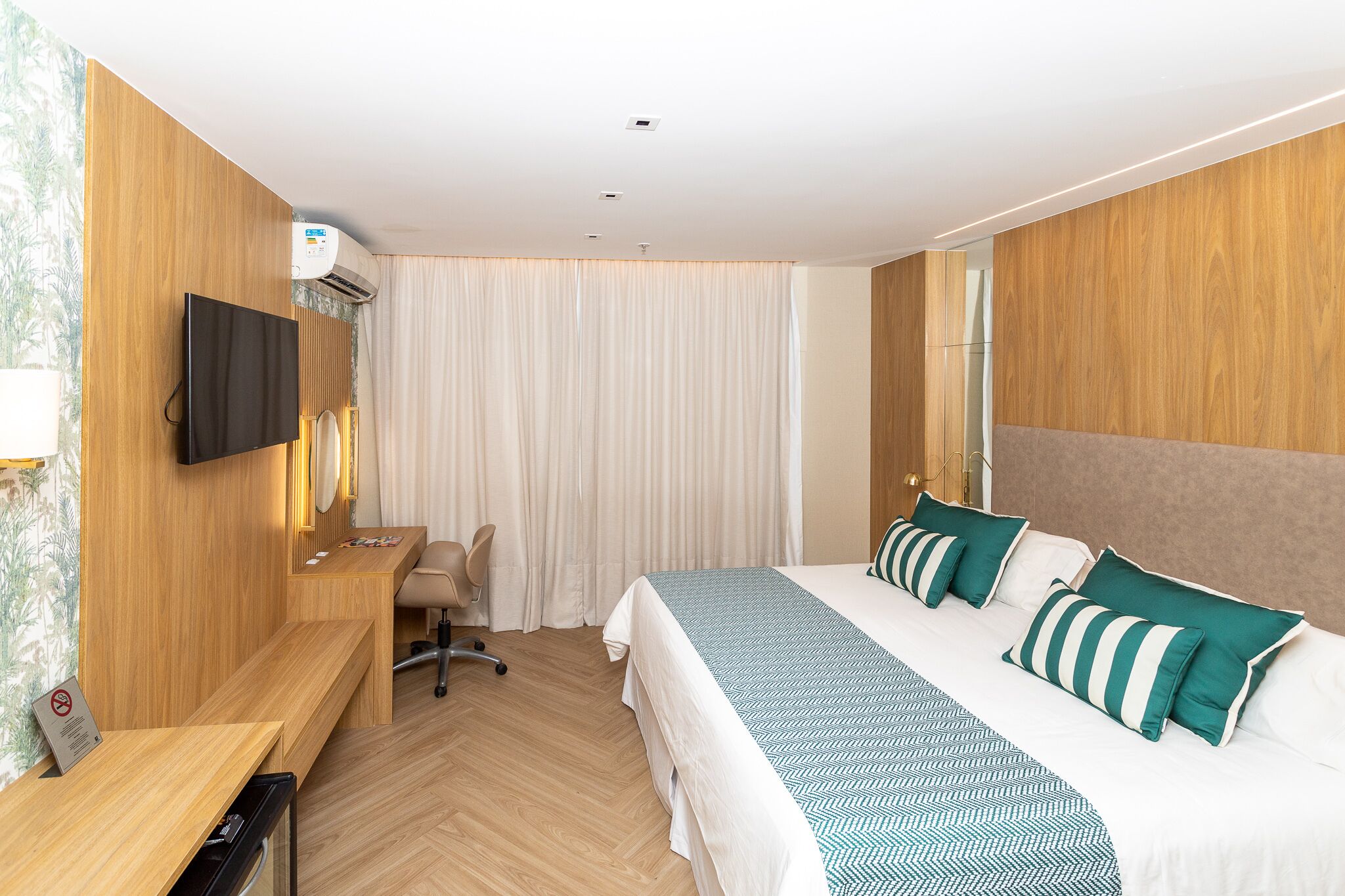 A hotel room with a wooden desk, a bed with green and white striped pillows, a television mounted on the wall, and an air conditioning unit.
